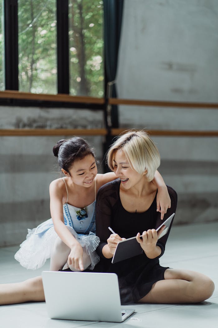 Cheerful ethnic girl embracing dance teacher writing information in notebook while pointing at laptop